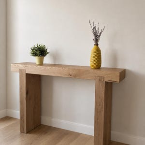 May include: A rectangular console table made of light brown wood with a thick top and two sturdy vertical legs. On the table sits a small green plant in a yellow pot and a yellow textured vase containing a sprig of dried lavender flowers.