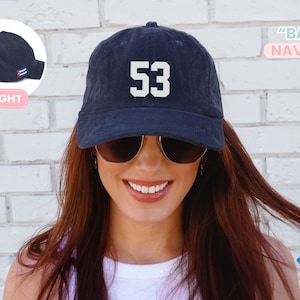 May include: Navy blue baseball cap with the number 53 in white block letters. The cap has a curved brim and an adjustable strap. The text "BA703" and "NAVY BLUE" are also visible. The cap is worn by a person.