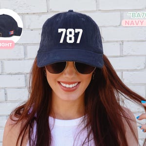 May include: Navy blue baseball cap with the number "787" in white block letters. The cap has a curved brim and an adjustable strap. The text "BA703" and "NAVY BLUE" are also visible.
