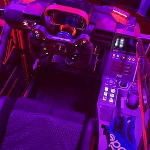 May include: A racing simulator cockpit with a steering wheel, dashboard, and seat. The cockpit is illuminated with purple and red lighting, and a pair of blue gloves are visible.