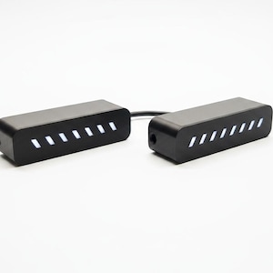 Può includere: Due componenti elettronici neri rettangolari collegati da un breve cavo nero. Ogni unità presenta una serie di sette fessure bianche inclinate e illuminate sulla parte anteriore e una porta circolare sul lato, con un design moderno e pulito.