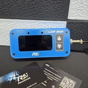 Może przedstawiać: Niebieskie urządzenie "LAP BOX" z ekranem i przyciskami, z logo "RS RaceSense". Zawiera dwie wizytówki z logo "RaceSense", stroną internetową i kodem QR. Urządzenie jest zaprojektowane i zbudowane dla Ciebie.