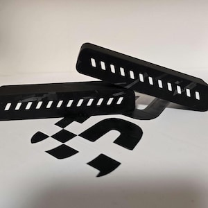 Puede incluir: Dos objetos rectangulares negros con una serie de recortes rectangulares blancos. Los objetos están colocados en ángulo, uno ligeramente por encima del otro. Un logotipo de bandera a cuadros en blanco y negro es visible debajo.