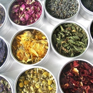 May include: Assortment of dried herbs and flowers in small metal tins. The tins are arranged in a circle on a white surface. The herbs and flowers include lavender, rose petals, mint leaves, chamomile, and blue flowers.