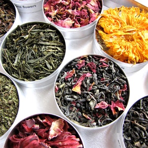 May include: Assortment of dried tea leaves and flowers in small, round metal tins. The teas include green tea, rose petals, and a vibrant orange flower. The tins are arranged in a circular pattern, offering a colorful display of herbal teas.