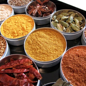 May include: A collection of spices in small metal containers. The spices include coriander seeds, turmeric, cumin, star anise, cardamom pods, and chili powder.