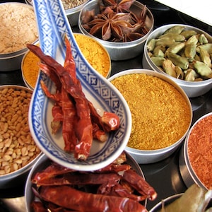 May include: A collection of spices in small metal bowls, including star anise, cardamom pods, dried chili peppers, turmeric, and cumin. The spices are arranged on a black surface with a white spoon.