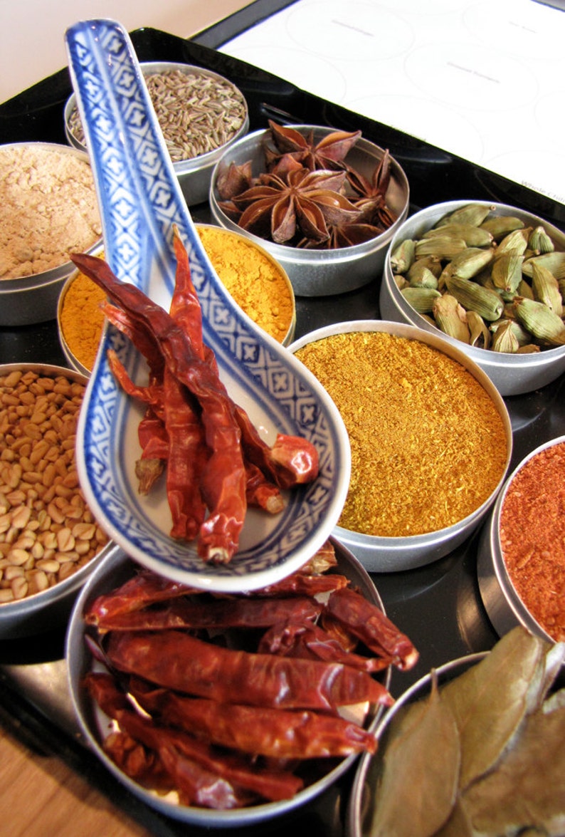 May include: A collection of spices in small metal bowls, including star anise, cardamom pods, dried chili peppers, turmeric, and cumin. The spices are arranged on a black surface with a white spoon.