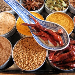 May include: A collection of spices in small metal containers, including star anise, cardamom pods, turmeric powder, cumin seeds, and dried red chili peppers. A blue and white ceramic spoon holds a few red chili peppers.