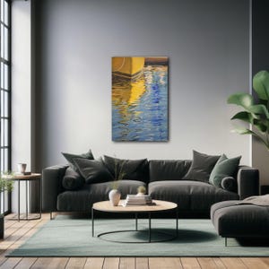 Op de afbeelding: Abstracte schilderij van een gele boot die wordt weerspiegeld in blauw water. Het schilderij is ingelijst en hangt aan een grijze muur boven een grijze bank.