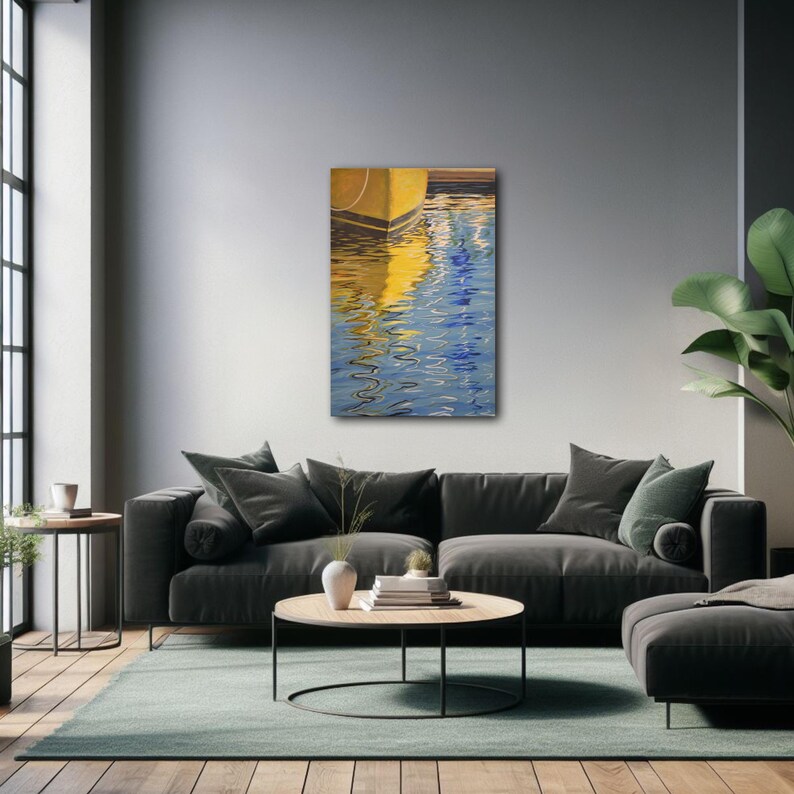 Op de afbeelding: Abstracte schilderij van een gele boot die wordt weerspiegeld in blauw water. Het schilderij is ingelijst en hangt aan een grijze muur boven een grijze bank.