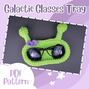 May include: A green crocheted glasses tray shaped like an alien with a purple bow. The tray has a black eyeglass holder and the text "PDF pattern" is visible in the image.