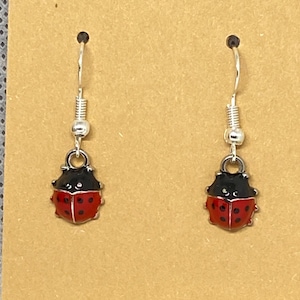 May include: A pair of drop earrings featuring small ladybird charms with glossy red wings, black spots, and black heads. The charms are attached to silver-toned metal fishhook wires with a small bead detail at the top of each earring hook.