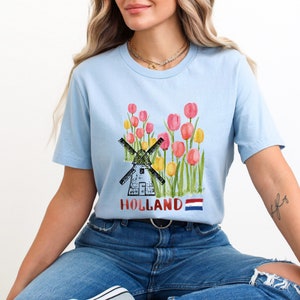 May include: A light blue t-shirt featuring a graphic of a windmill and tulips. The text "HOLLAND" is printed on the shirt, along with a small Dutch flag.