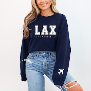 May include: A navy blue cropped sweatshirt with white text that reads "LAX LOS ANGELES, CA" and a white airplane graphic on the sleeve.