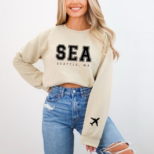 May include: A beige cropped sweatshirt with black text that reads "SEA SEATTLE, WA" and a black airplane silhouette on the left sleeve.