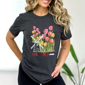 May include: A dark grey t-shirt featuring a white windmill and a field of pink and yellow tulips. The text "HOLLAND" is printed on the shirt with a small Dutch flag.