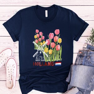 May include: A navy blue t-shirt featuring a watercolour illustration of a windmill and tulips. The text "HOLLAND" is printed on the shirt, along with the Dutch flag.