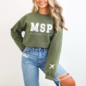 Puede incluir: Una sudadera verde con texto blanco que dice "MSP Minneapolis, MN" y un gráfico de avión blanco en la manga.