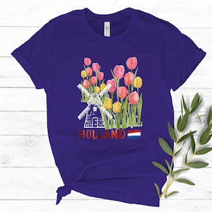 May include: Purple t-shirt featuring a white windmill and red, yellow, and pink tulips. The shirt also has the word "HOLLAND" and the Dutch flag printed on it.