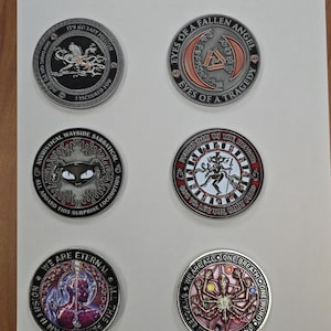 Puede incluir: Seis monedas de metal detalladas con diseños y texto intrincados. Las monedas presentan varios temas, incluyendo un ángel caído, una araña y otras imágenes simbólicas. Las monedas se exhiben sobre una superficie blanca.