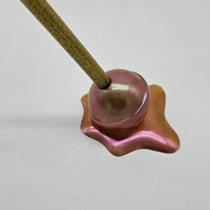 May include: A gold glitter pen with a pink and gold iridescent lollipop-shaped base. The base is melted and dripping.