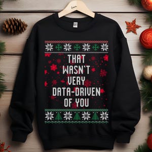 Könnte beinhalten: Schwarzer Pullover mit dem weißen Text "THAT WASN'T VERY DATA-DRIVEN OF YOU". Das Design umfasst Schneeflocken und Weihnachtsbäume in Grün und Weiß, passend für die Weihnachtszeit.