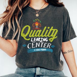 May include: Dark gray t-shirt with a distressed look. The shirt features the words "Quality Learning Center" in green and white, with a treehouse graphic and the phone number 1-800-FRAUD in blue. The shirt is a casual wear item.