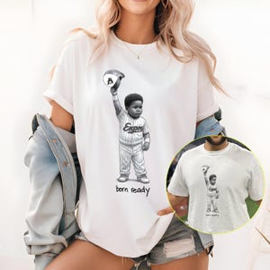 Peut inclure: T-shirt blanc avec une illustration en noir et blanc d'un jeune joueur de baseball levant une casquette. Le texte "born ready" est en dessous. Le joueur porte un uniforme de baseball.