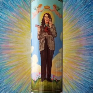 May include: A glass candle holder with a white candle inside. The candle holder features a colorful image of a person holding a guitar with two angels above them. The image has a green border and a blue background.