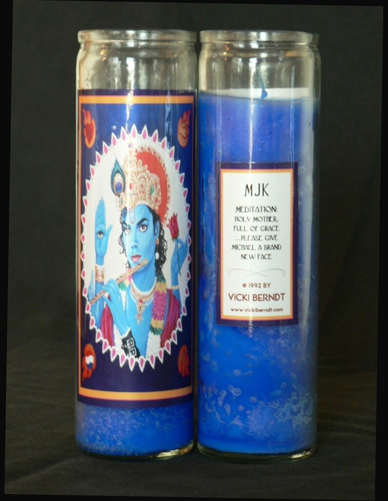 May include: Two blue glass candle jars with a blue wax interior. The left jar has a label with an image of a blue-skinned deity playing a flute. The label text reads "MJK Meditation: Holy Mother, Full of Grace... Please give Michael a brand new face. &copy; 1992 by Vicki Berndt www.vickiberndt.com".