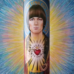 May include: A blue glass candle with a red and gold label featuring a portrait of a man with brown hair and a red heart with a dagger. The label reads "Greetings Go Forth" and "Saint Raymond Dee Ray" in a gold font.