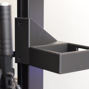 May include: A black, rectangular shelf attached to a vertical support structure. The shelf is open-topped and appears to be made of a matte material. The support structure has a series of vertical bars and a cylindrical component.