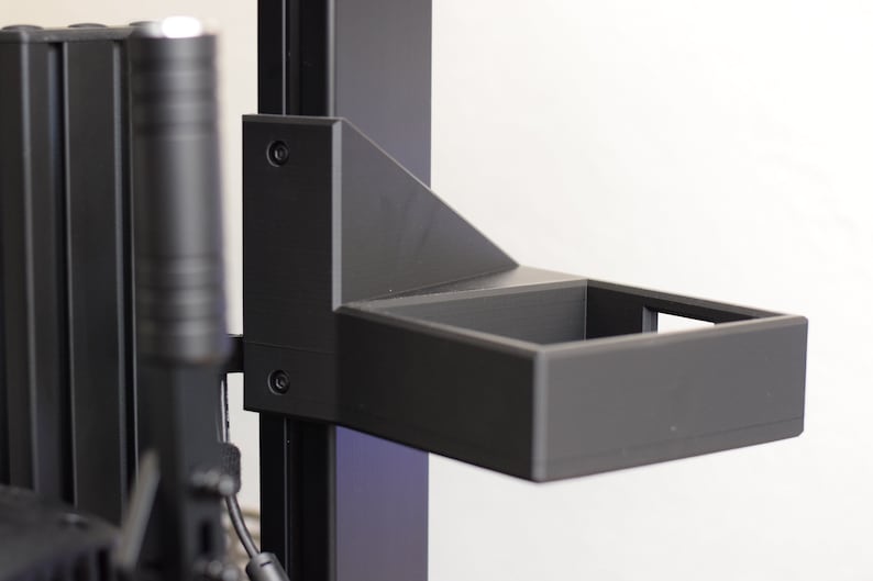 May include: A black, rectangular shelf attached to a vertical support structure. The shelf is open-topped and appears to be made of a matte material. The support structure has a series of vertical bars and a cylindrical component.