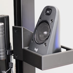 May include: A black Logitech speaker is mounted on a black metal bracket. The speaker has a mesh grill and the Logitech logo. The bracket is attached to a black vertical support.
