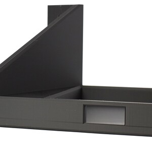 May include: A dark gray, angled storage box with a rectangular compartment and a smaller, recessed section. The design includes a sloped back and a flat base, with a small screw visible on the left. The box is made of a matte material.