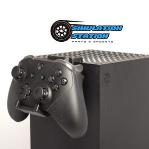 May include: A black Xbox Series X console with a black Xbox controller mounted on the side. The controller is attached to the console with a black bracket. The console has a textured surface and the controller has a textured grip.