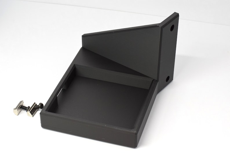 May include: A black, geometric wall-mounted shelf with a stepped design. The shelf has a rectangular base, a middle shelf, and an angled top section. Two silver cufflinks are placed near the shelf.