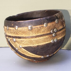 May include: A dark brown, rounded bowl featuring a textured, light-colored carved pattern around its exterior. The bowl is decorated with numerous small, circular, silver-toned metal studs embedded into the surface along the carved lines.
