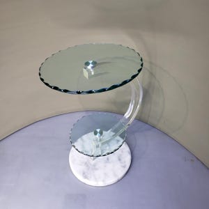 May include: A two-tier side table featuring a circular white marble base, a curved clear acrylic support structure, and two round glass shelves with scalloped edges. Each glass shelf is secured to the acrylic frame with a silver metal fastener.