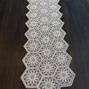 May include: Cream-colored, open-weave table runner with a floral design. The runner is made of hexagonal shapes, each featuring a flower. The edges are scalloped, and the runner is placed on a dark wooden surface.