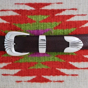 Puede incluir: Un cinturón de cuero marrón con una hebilla plateada. La hebilla tiene un diseño decorativo con un borde festoneado. El cinturón está sobre un fondo estampado blanco y rojo.
