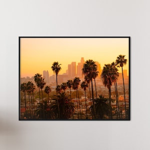 Puede incluir: Una fotografía enmarcada de una ciudad con palmeras en primer plano. La ciudad se recorta contra un cielo de puesta de sol naranja brillante.