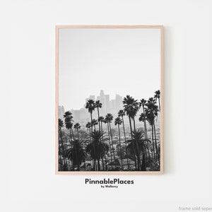 May include: Black and white photograph of a city skyline with palm trees in the foreground. The city is in the distance and the palm trees are in the foreground. The image is framed in a light wood frame. The text "PinnablePlaces by Wallency" is at the bottom of the image.
