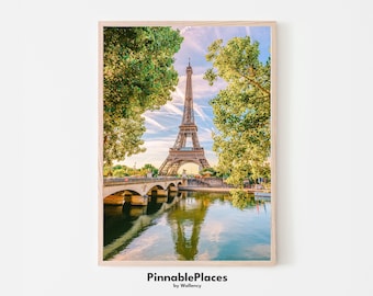 May include: A photograph of the Eiffel Tower in Paris, France. The tower is seen from across the River Seine, with green trees on either side of the river. The water reflects the tower and the sky, which is a light blue with white clouds.