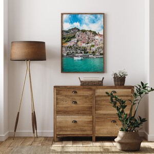 May include: A framed photograph of a colorful village built into a cliffside overlooking a turquoise body of water. The photo is displayed on a wooden dresser with two drawers. A potted plant and a basket of books are also on the dresser.