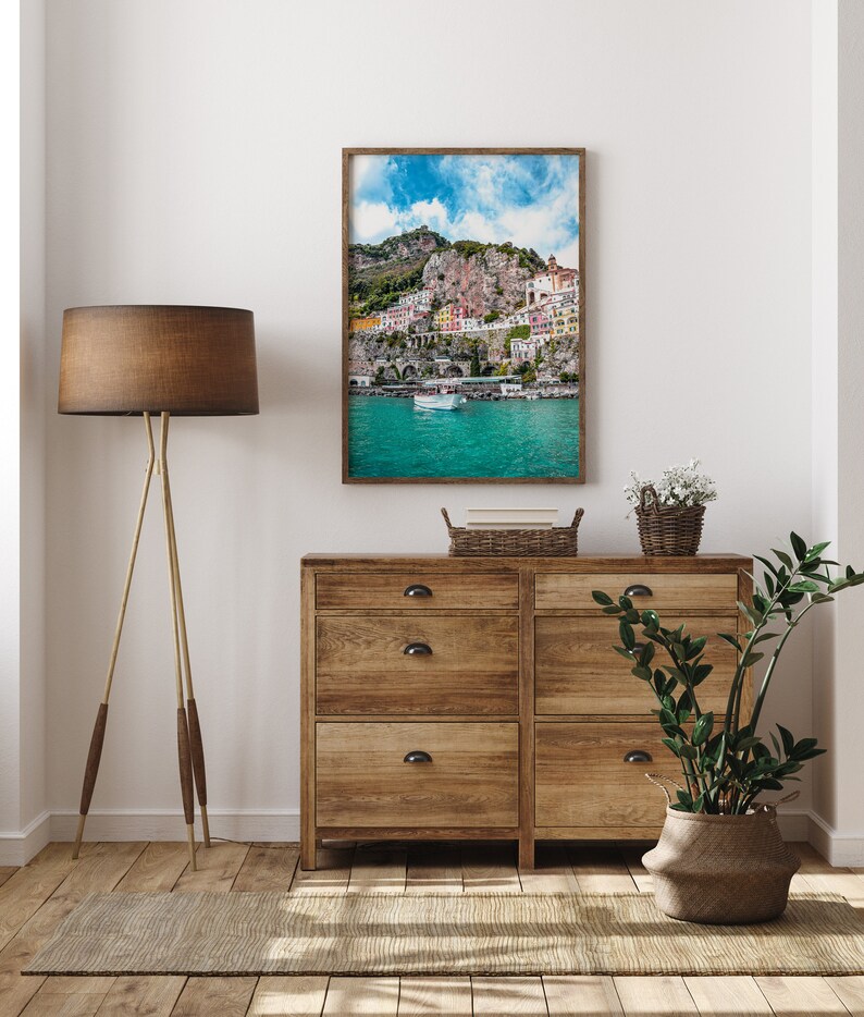 May include: A framed photograph of a colorful village built into a cliffside overlooking a turquoise body of water. The photo is displayed on a wooden dresser with two drawers. A potted plant and a basket of books are also on the dresser.