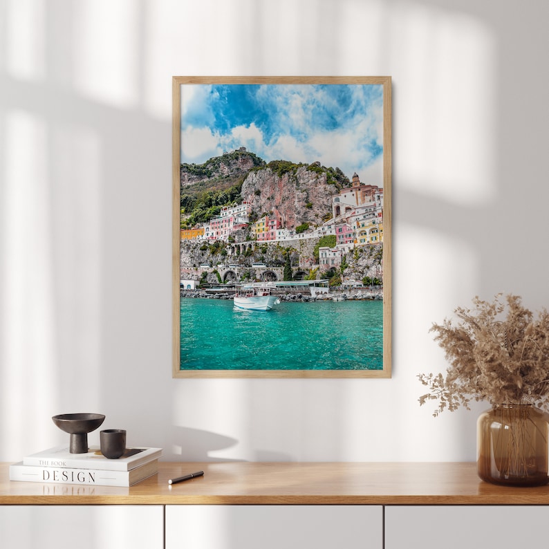 May include: A framed photograph of a colorful town built into a cliffside overlooking a turquoise body of water. A white boat is docked in the water.
