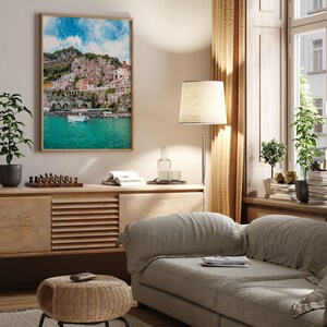 May include: A living room interior with a light gray sofa, a wooden cabinet, a wicker ottoman, a floor lamp, and a framed print of a colorful Italian coastal town.