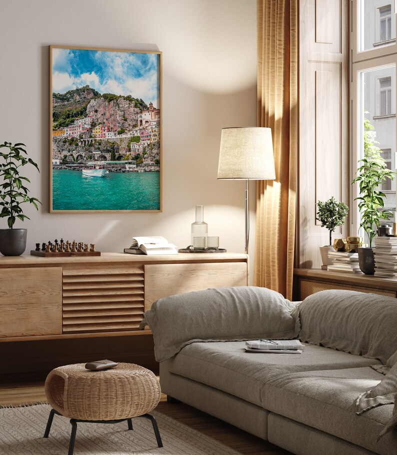 May include: A living room interior with a light gray sofa, a wooden cabinet, a wicker ottoman, a floor lamp, and a framed print of a colorful Italian coastal town.